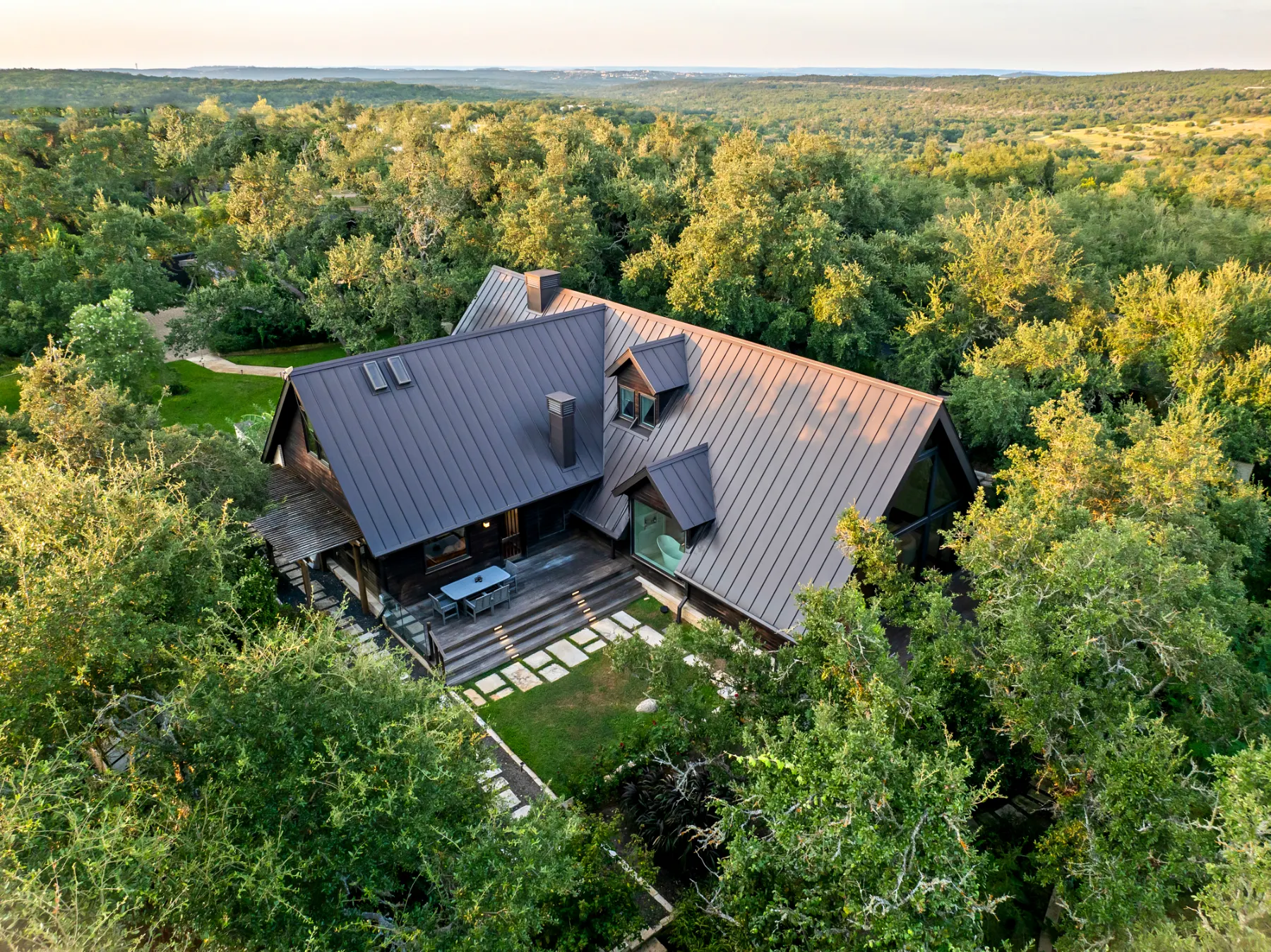 Aerial view of a dark-roofed house surrounded by dense trees, with Hill Country stretching to the horizon