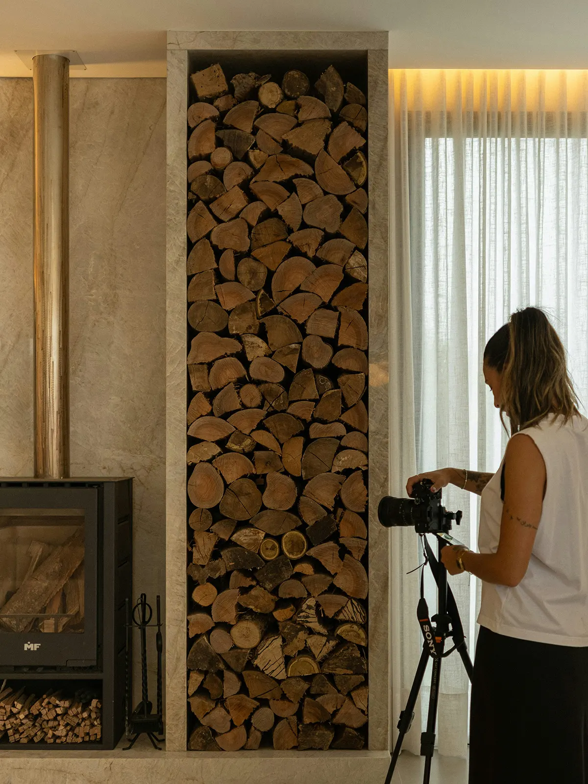 A photographer with a camera on a tripod shooting a living room with a wood stove and a stacked log wall