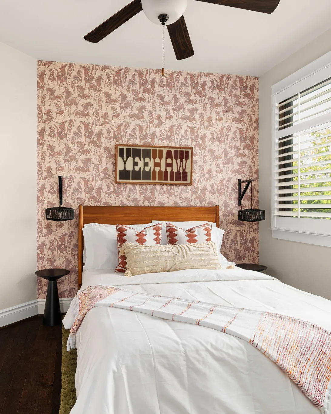 A bedroom with pink botanical wallpaper, a "Yeehaw" sign above the bed and a striped throw