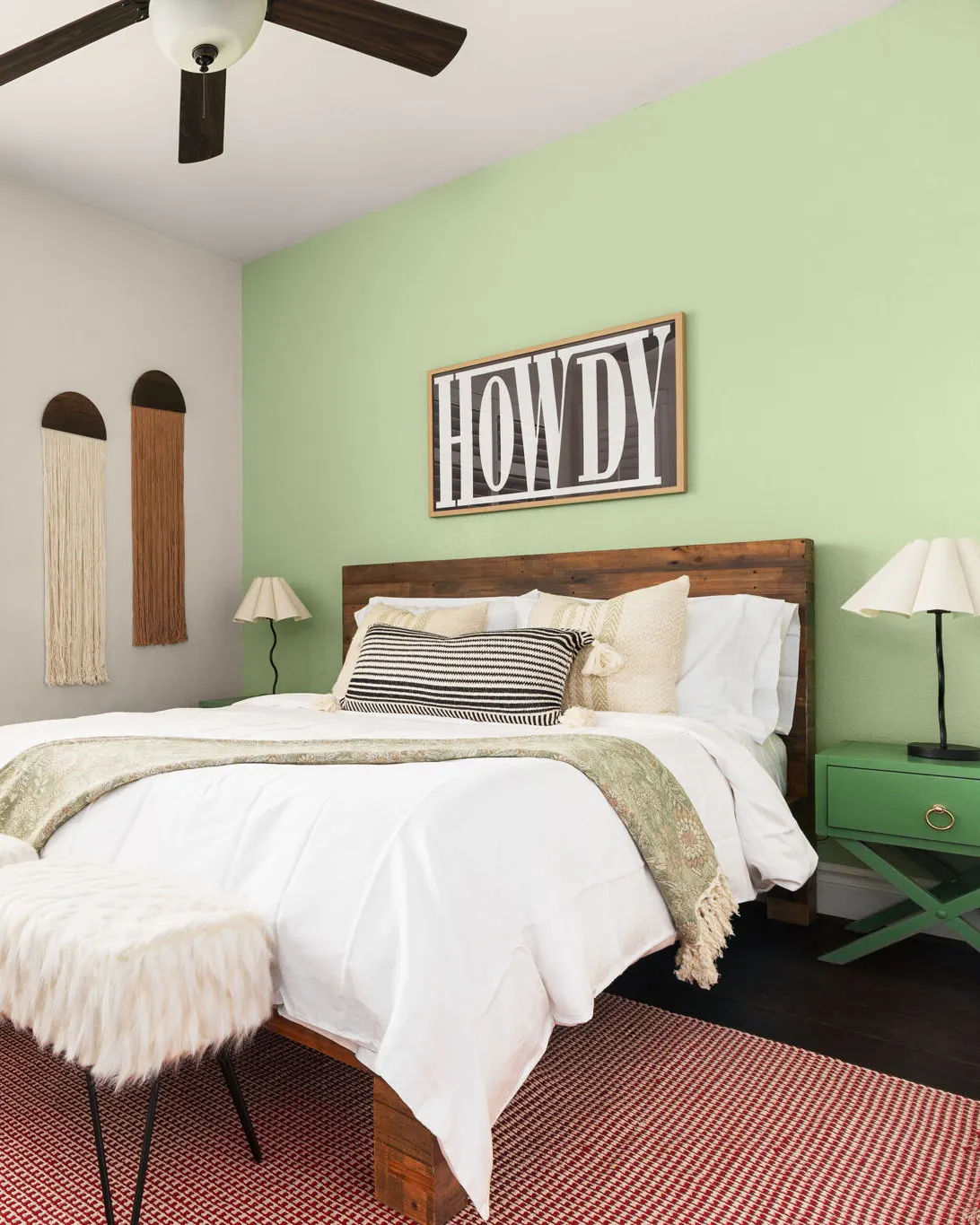A bedroom with a sage green accent wall, a "Howdy" sign above the bed and a red striped rug