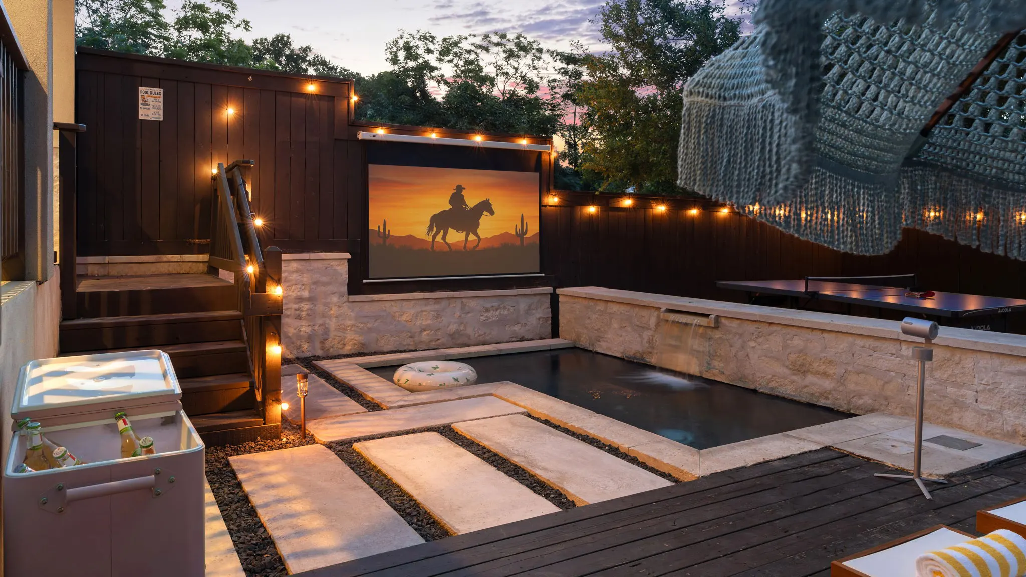 A cold plunge pool and stone steps at dusk under string lights, facing an outdoor screen showing a cowboy on horseback