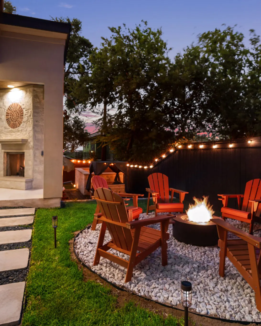 Red Adirondack chairs around a fire pit on a white gravel circle, with string lights along the fence at dusk