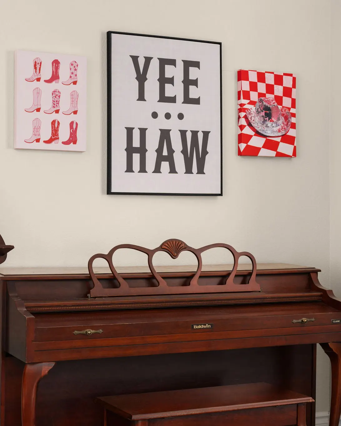 An upright wooden piano beneath framed pink cowboy-boot and "Yee Haw" prints