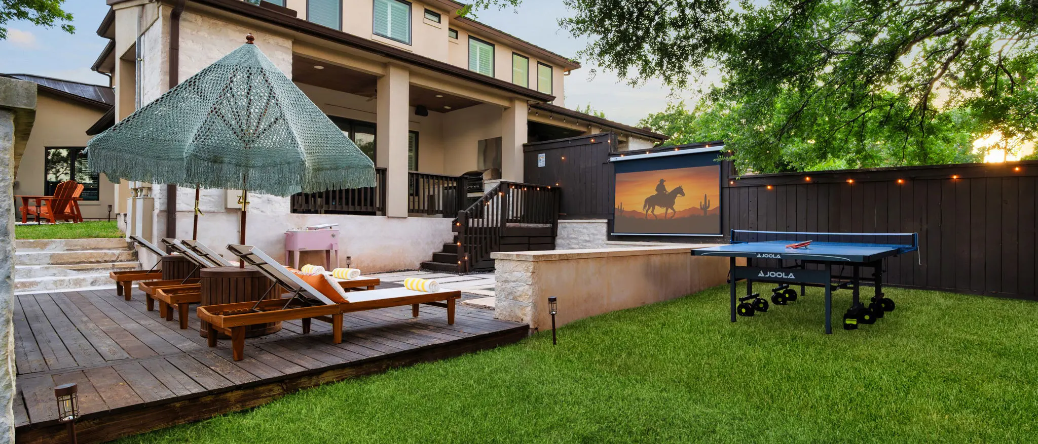 The Sunset Saloon backyard at dusk: a wooden deck with loungers under a fringed umbrella, a ping-pong table, and an outdoor movie screen on the fence