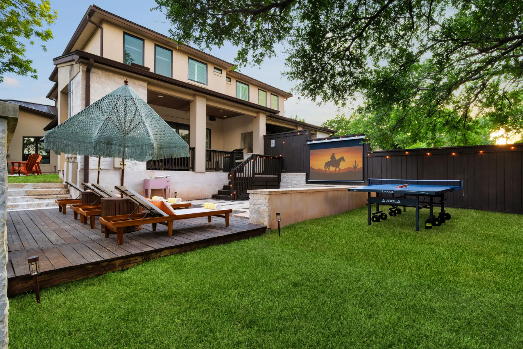 The backyard after the redesign: a deck with loungers under a fringed umbrella, a ping-pong table, and an outdoor movie screen at dusk